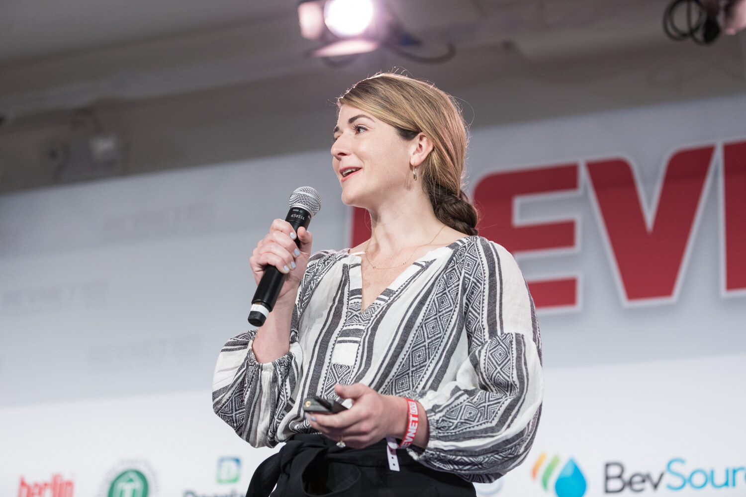 Woman in a black and white patterned blouse and low braid speaking into a microphone in front of BevSource and Berlin Packaging signs.