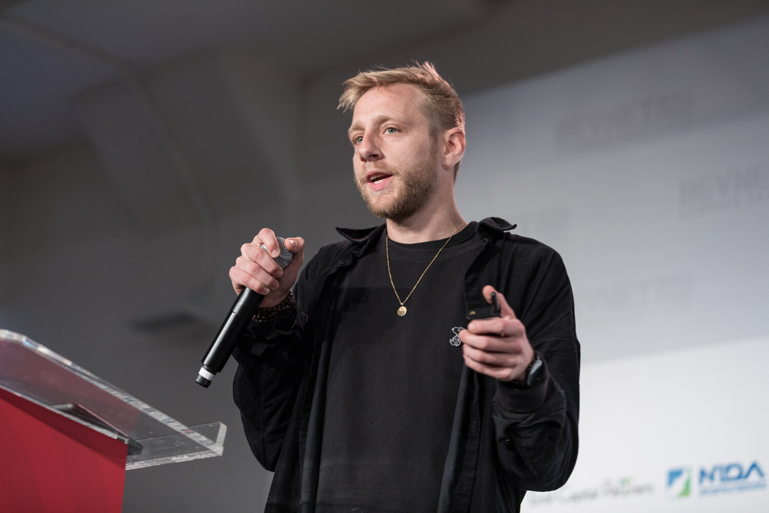 Man in a black overshirt and gold pendant holding a microphone beside the red podium.