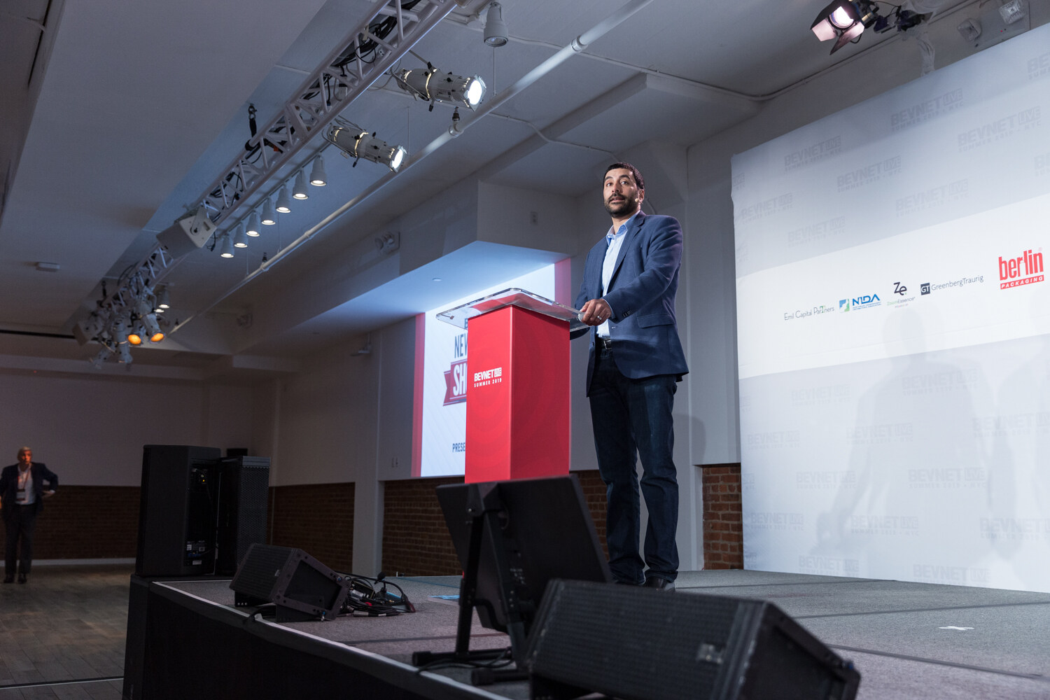 Man in a blue blazer standing at the red podium on the New Beverage Showdown stage before the pitches.