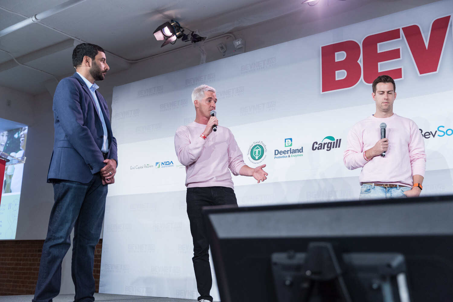 Man in a navy blazer and two men in pink sweatshirts standing on stage in front of the BevNET Live sign.