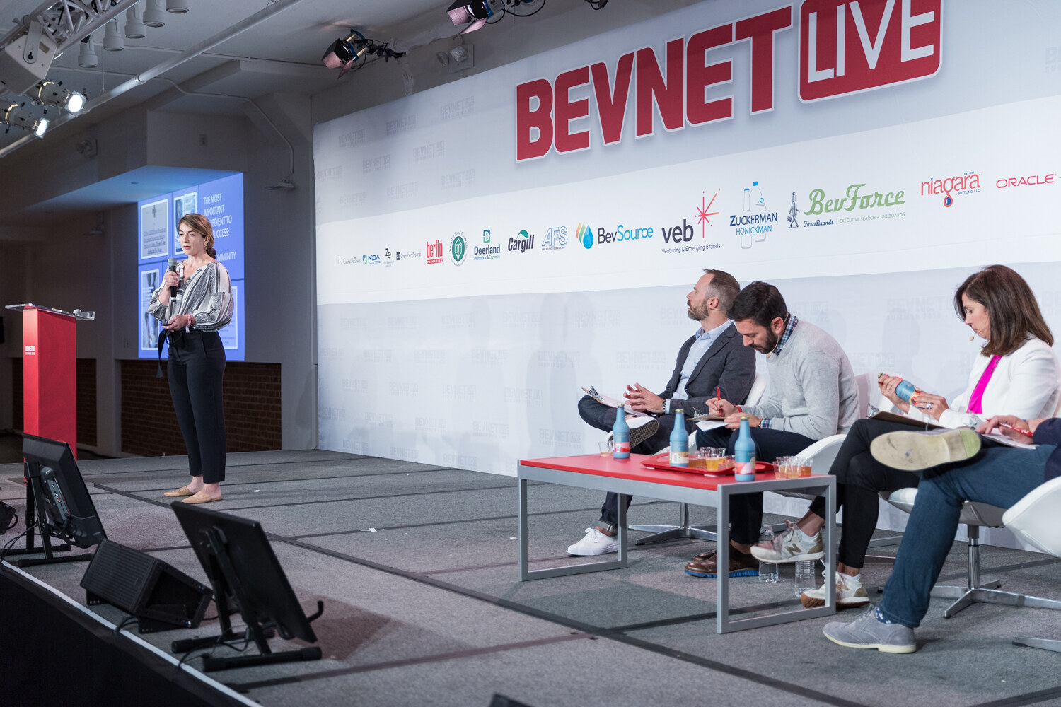 Woman presenting at the left of the stage while four judges sit at a red table.