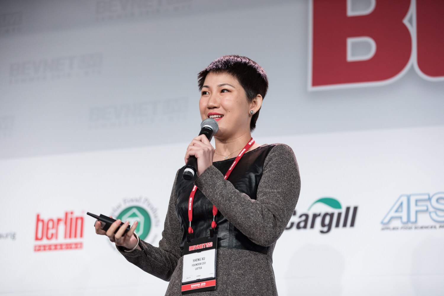 Woman in a gray dress with a black leather panel, a red lanyard and a name badge holding a clicker and microphone on stage.