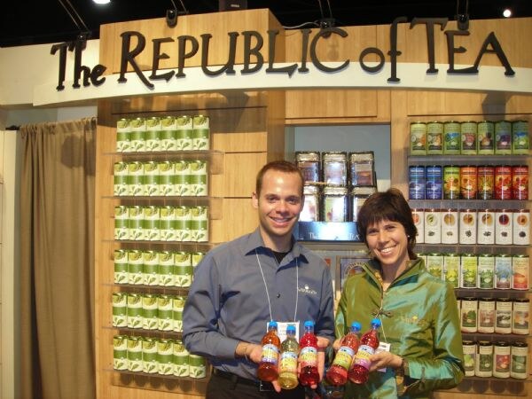 A man in a gray shirt and a woman in a green jacket each hold bottled teas in front of The Republic of Tea booth, with shelves of tea tins behind them.