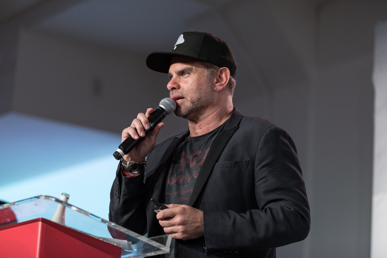 Man in a black cap and dark blazer speaking into a microphone at the red podium.