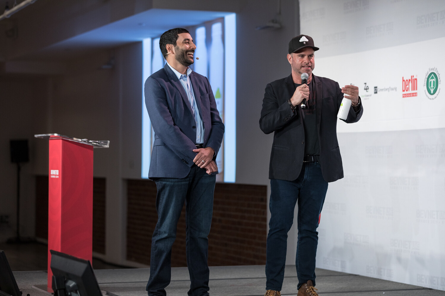 Man in a navy blazer and a man in a black cap holding a white bottle and microphone standing beside the red podium.
