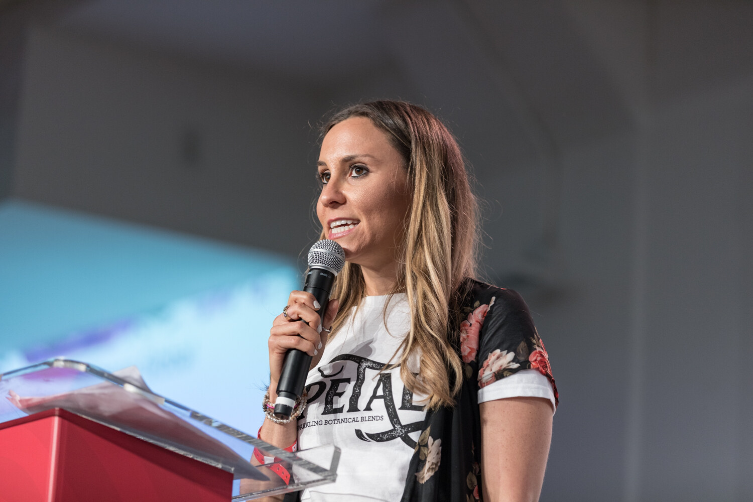 Woman in a Petal t-shirt and floral duster speaking into a microphone at the podium.