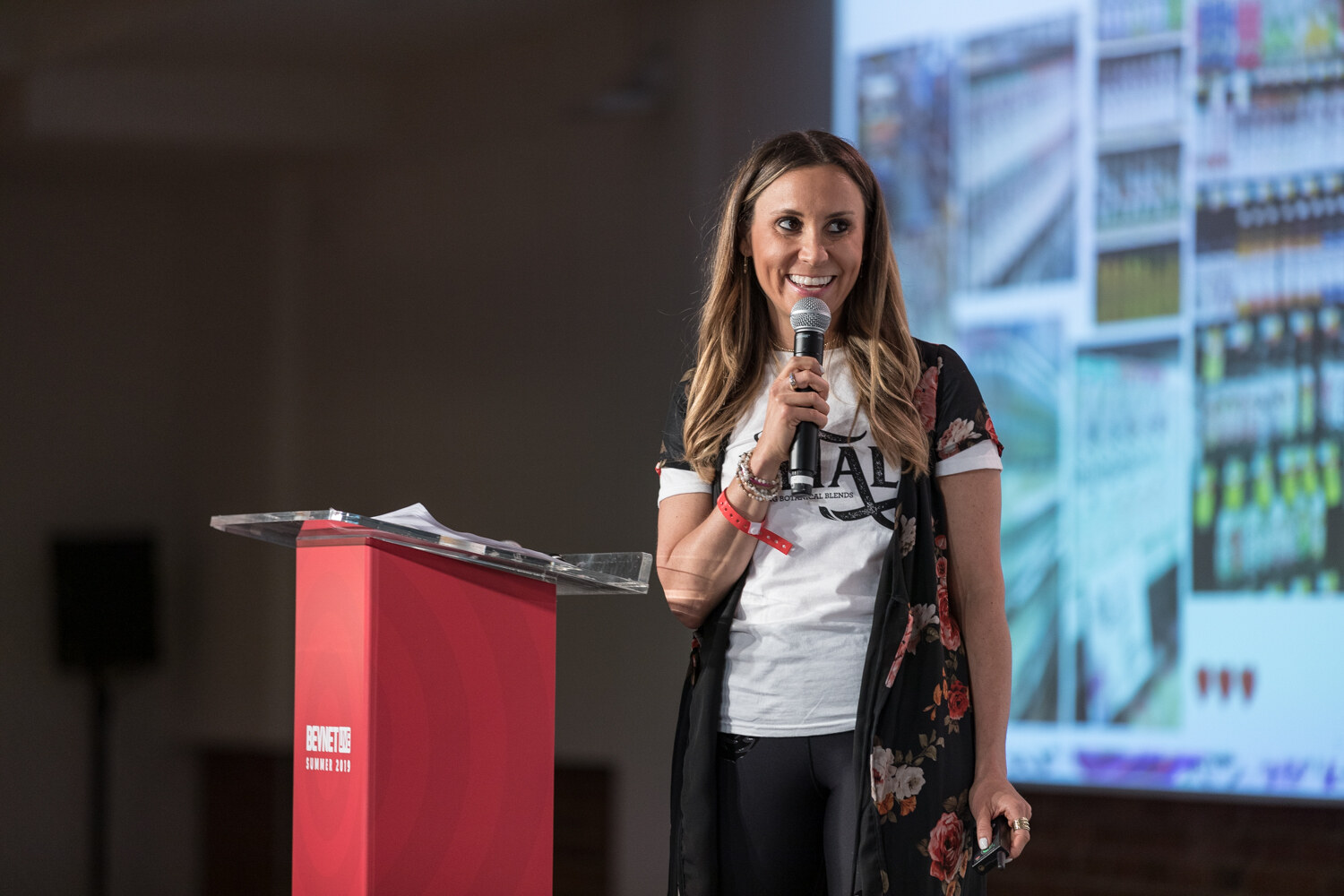 Smiling woman in a Petal t-shirt beside the red podium with a store shelf image on the screen behind her.