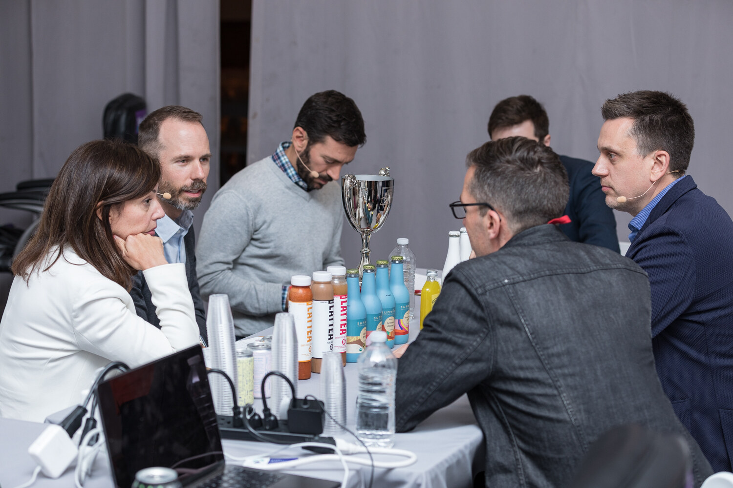 Judges around a table with a silver trophy and rows of beverage samples during deliberation.