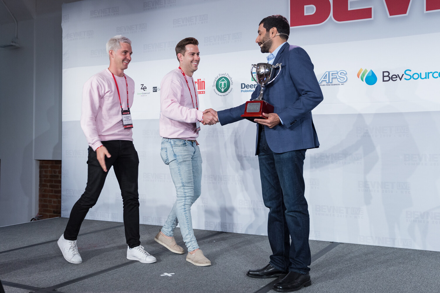 Winners in pink sweatshirts shaking hands with the host as they receive a silver trophy on stage.