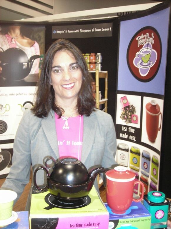 A smiling woman with dark hair in a gray blazer and pink shirt stands at The Tea Spot booth behind a black teapot, a red mug and boxes, with a purple Tea Spot banner beside her.