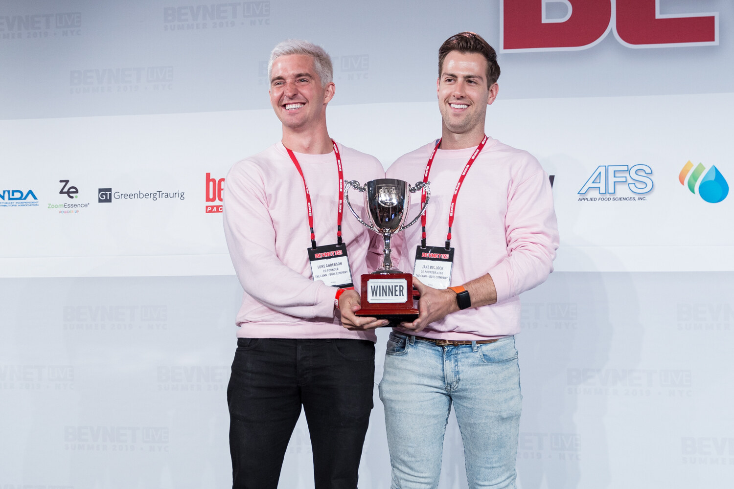 Two winners in pink sweatshirts holding up a silver trophy labeled Winner.