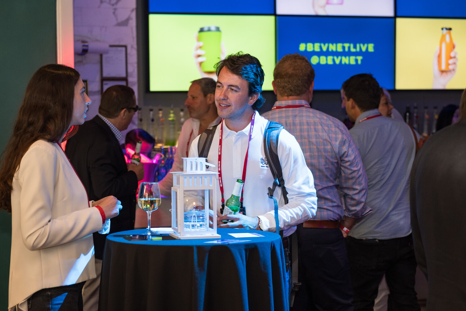 Man in a white shirt and lanyard smiling beside a lantern on a high table, beneath screens reading #BEVNETLIVE @BEVNET.