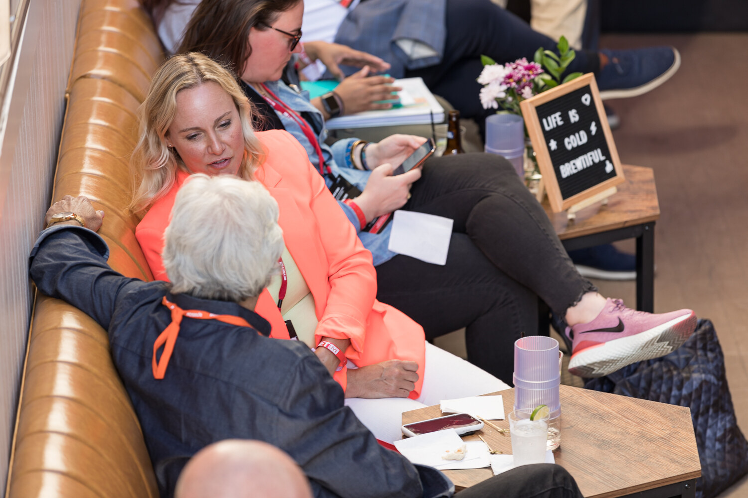 Women seated on a leather banquette beside a letterboard sign reading Life is cold brewtiful.
