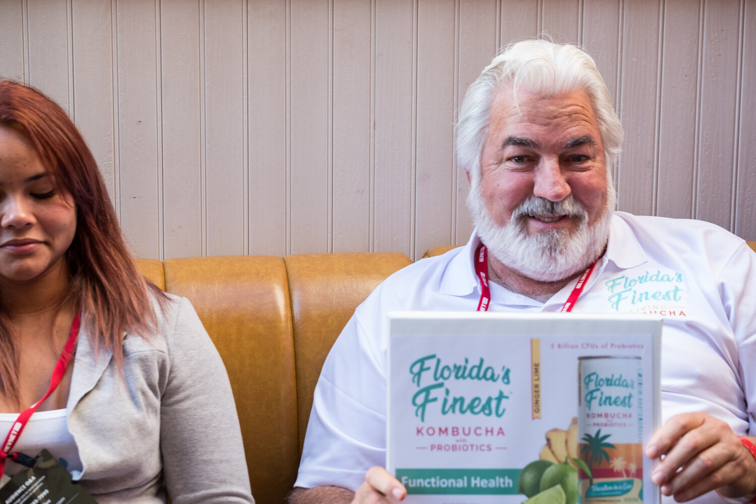 Bearded man in a white Florida's Finest Kombucha shirt holding up a brand sign in a leather booth, with a woman beside him.