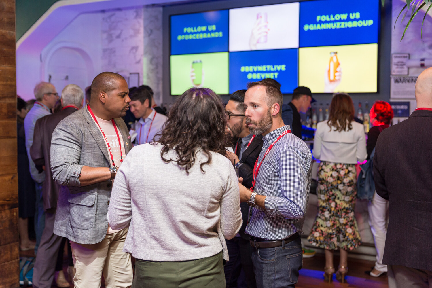 Attendees talking near the bar beneath screens showing #BEVNETLIVE and sponsor messages.