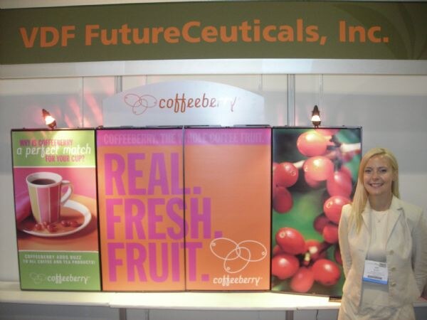 A smiling blonde woman with a name badge stands beside orange and pink Coffeeberry display panels reading Real. Fresh. Fruit., under a VDF FutureCeuticals, Inc. sign.