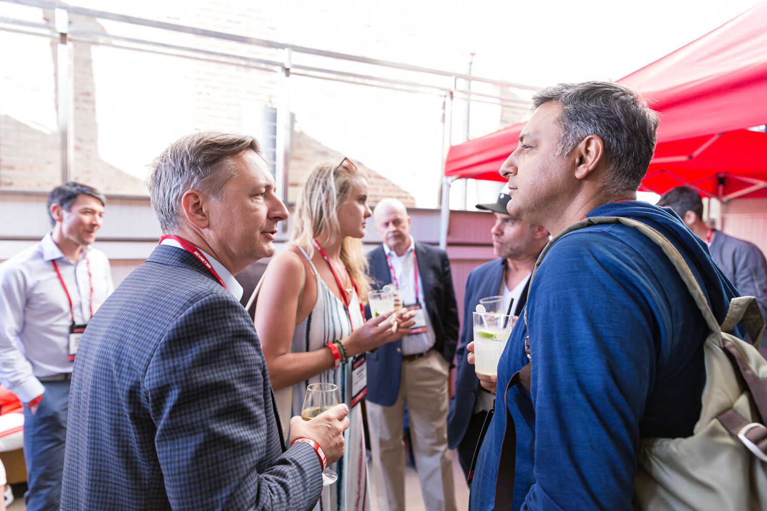 Attendees in blazers chatting with drinks on the rooftop terrace beneath a red canopy.
