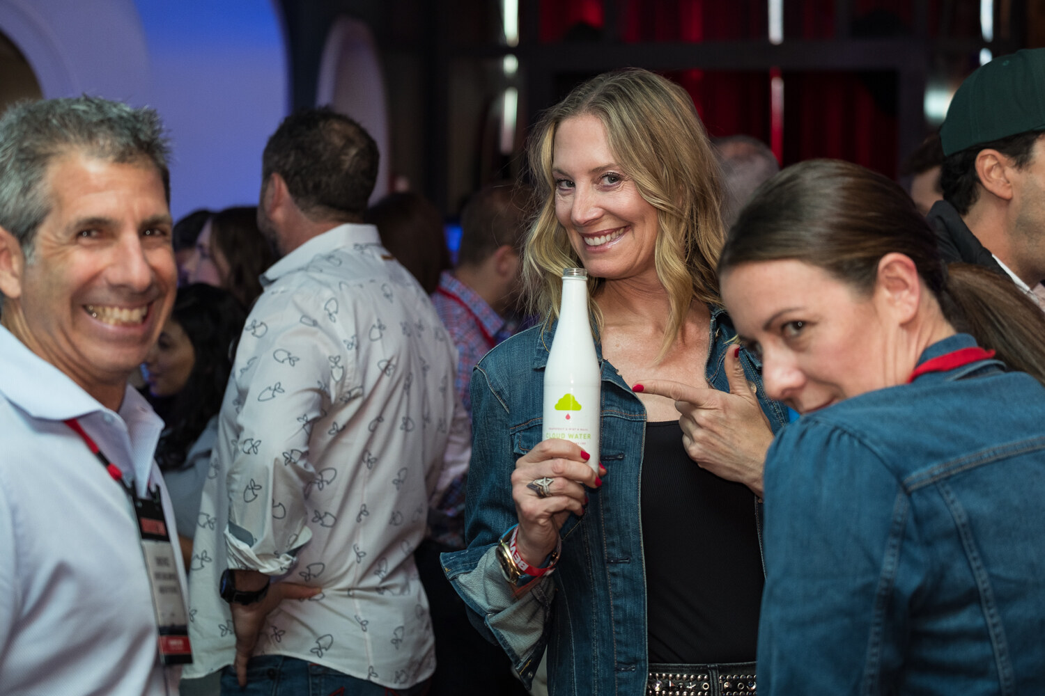 Woman in a denim jacket holding a bottle and smiling while another woman points at it, with a man smiling at left.