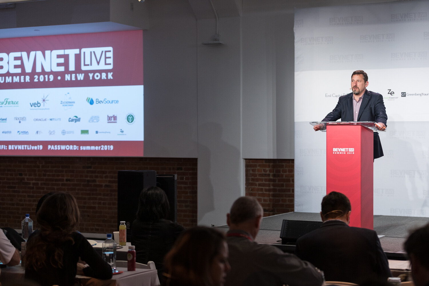 Host at the podium beside a screen reading BevNET Live Summer 2019 New York with the Wi-Fi details.