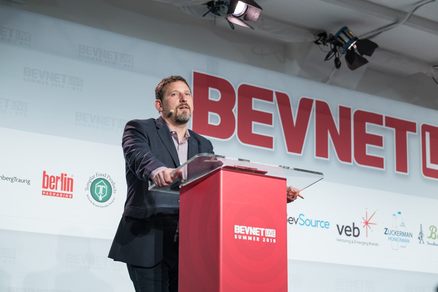 Presenter in a dark blazer leaning on the red BevNET Live podium during the welcome remarks.