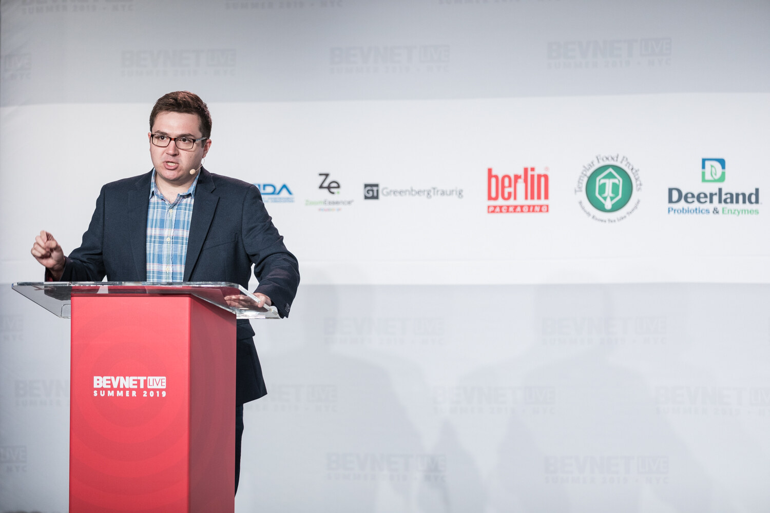 Presenter in a navy blazer and plaid shirt at the red BevNET Live podium with sponsor logos on the wall behind him.