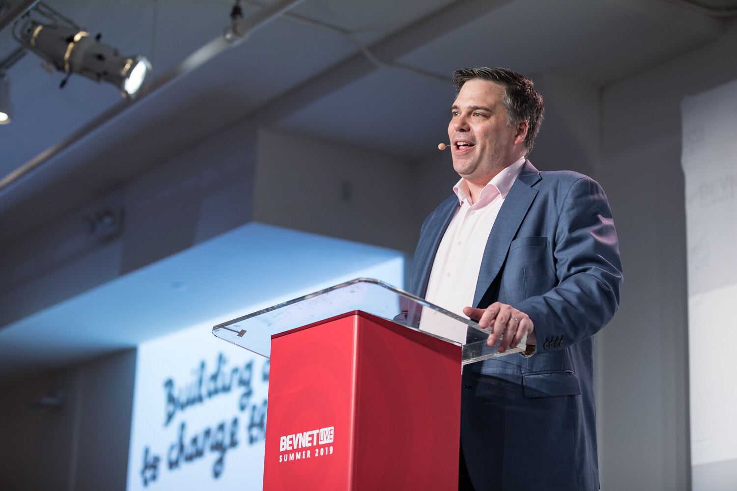 Man in a blue blazer smiling at the red podium with a slide reading Building to change behind him.
