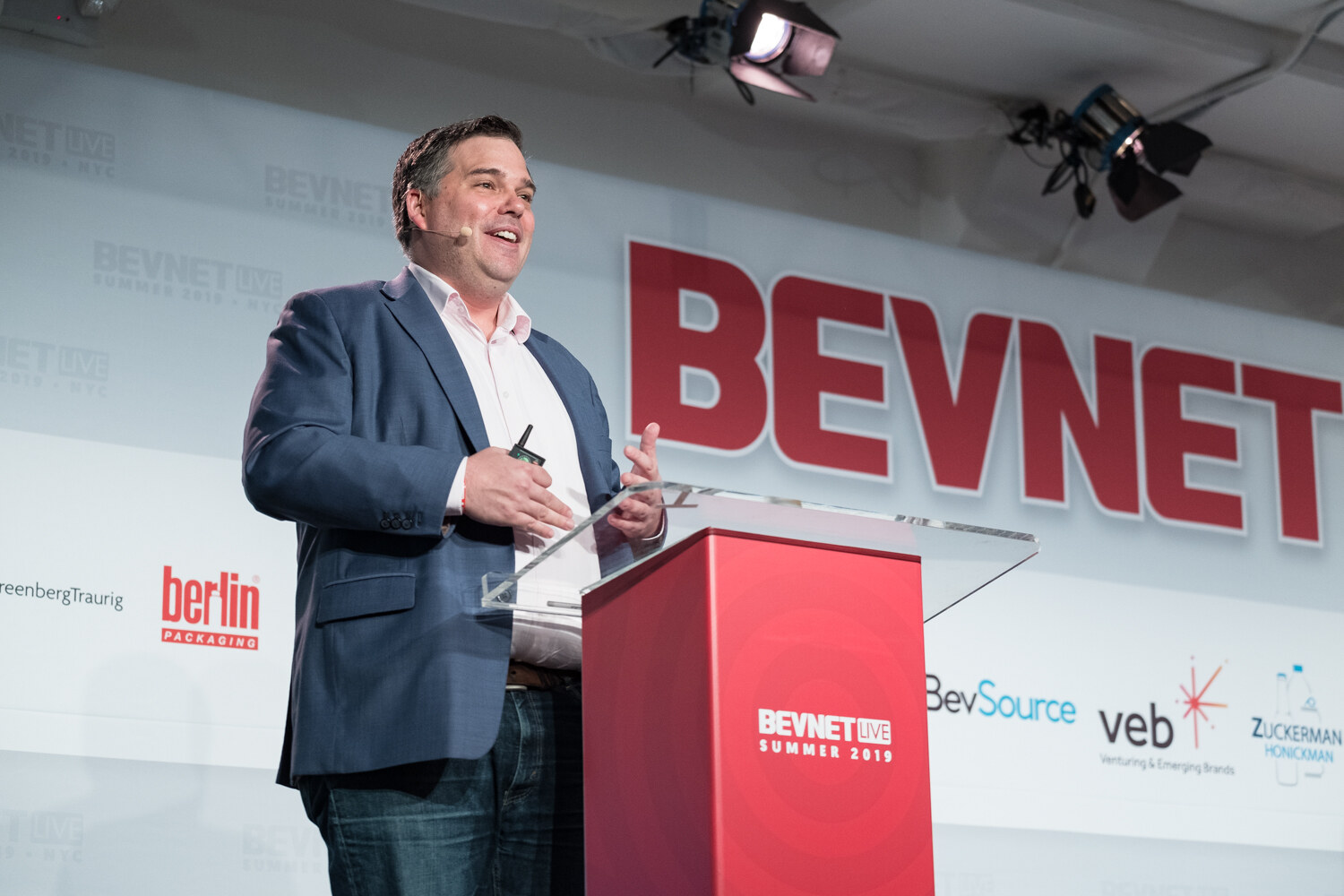 Man in a blue blazer smiling and gesturing at the red podium in front of the BEVNET sign and BevSource and veb logos.