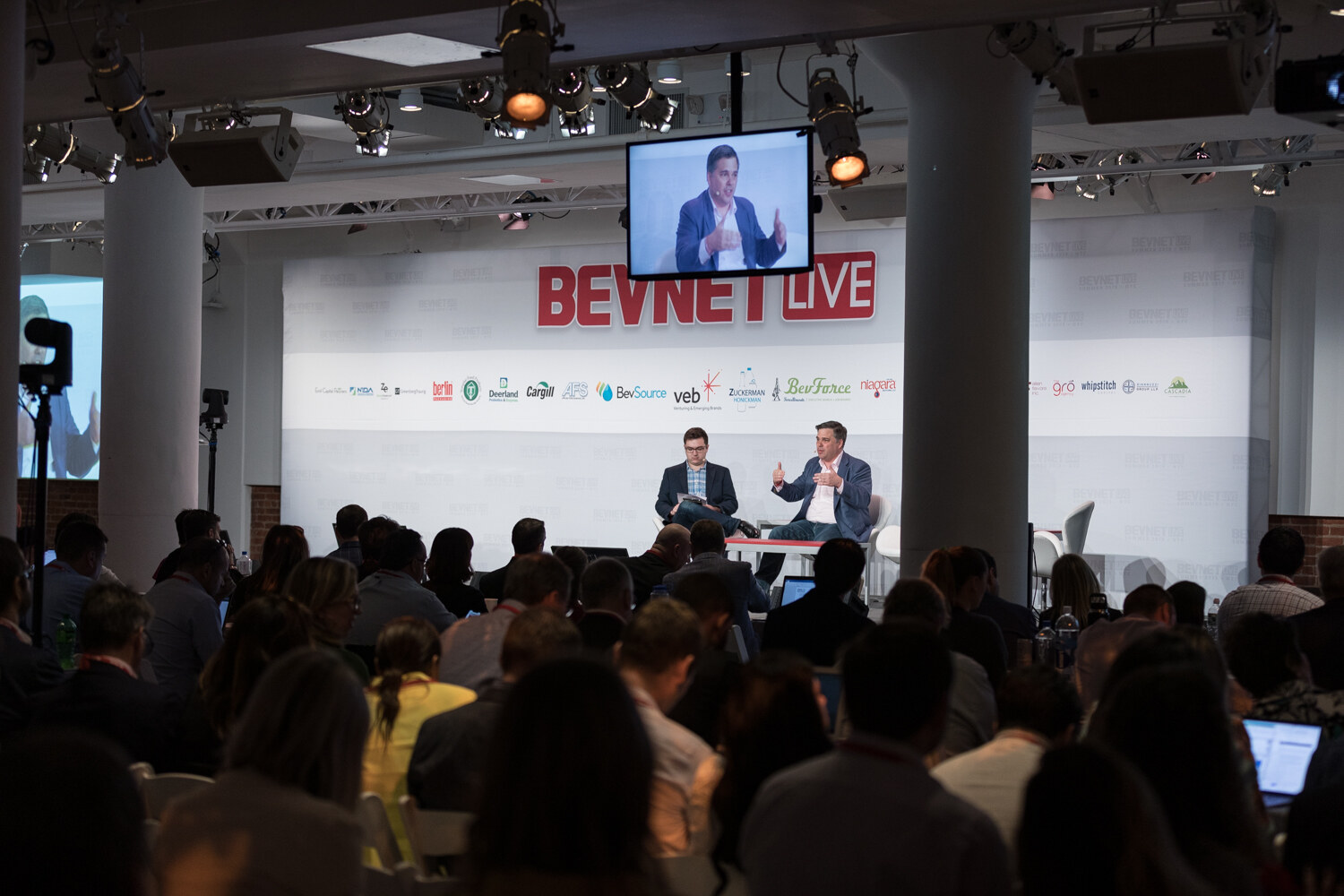 Wide view of the fireside chat with a video screen above the stage and a full audience in the foreground.