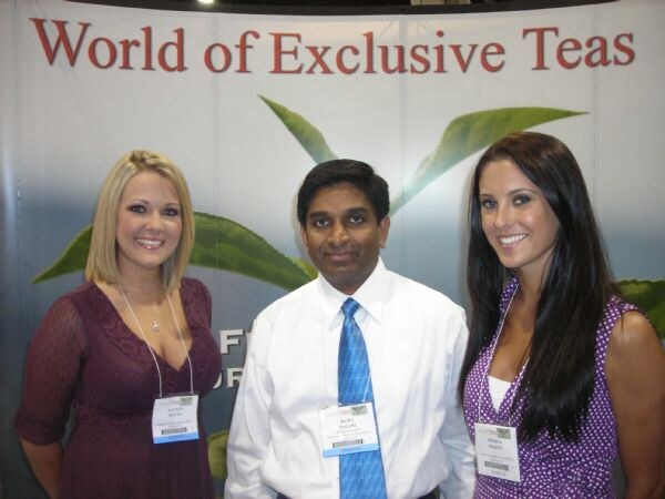 Two women and a man in a white shirt and blue tie, all wearing name badges, smile in front of a World of Exclusive Teas backdrop with green tea leaves.
