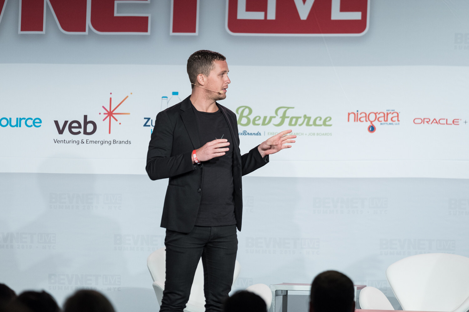 Speaker in a black blazer gesturing on stage in front of the BevNET Live sign.