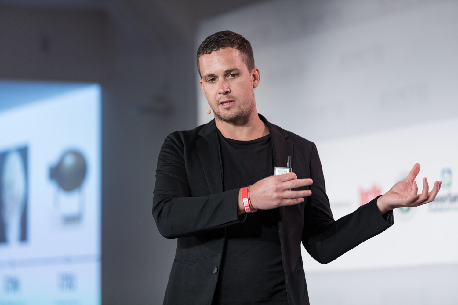 Speaker in a black blazer gesturing with an open hand in front of a slide of portraits.