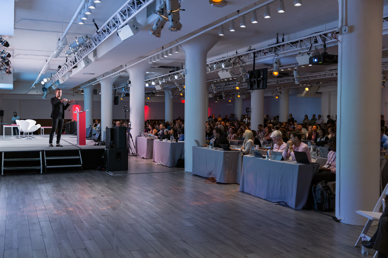 Wide view of the main session room from the floor, with a speaker on stage and a full audience at draped tables beneath white columns.