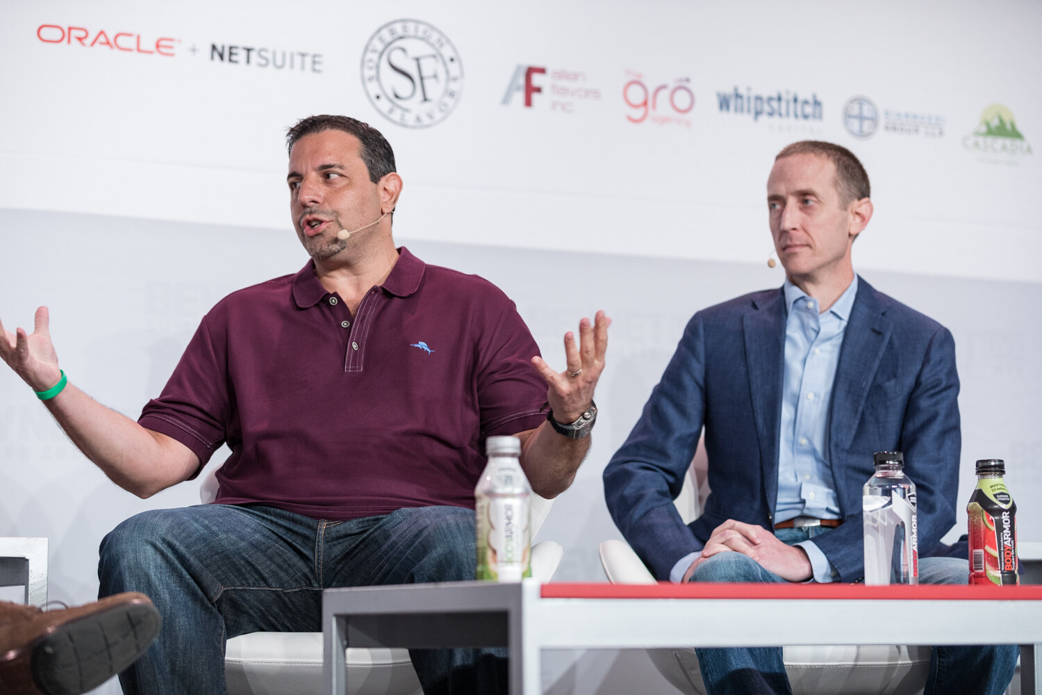 Man in a maroon polo gesturing with both hands beside a man in a navy blazer.