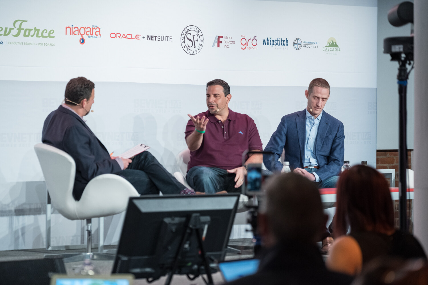 Interviewer in a dark blazer and two men in a maroon polo and a blue blazer seated on the BevNET Live stage with a camera and monitor in the foreground.