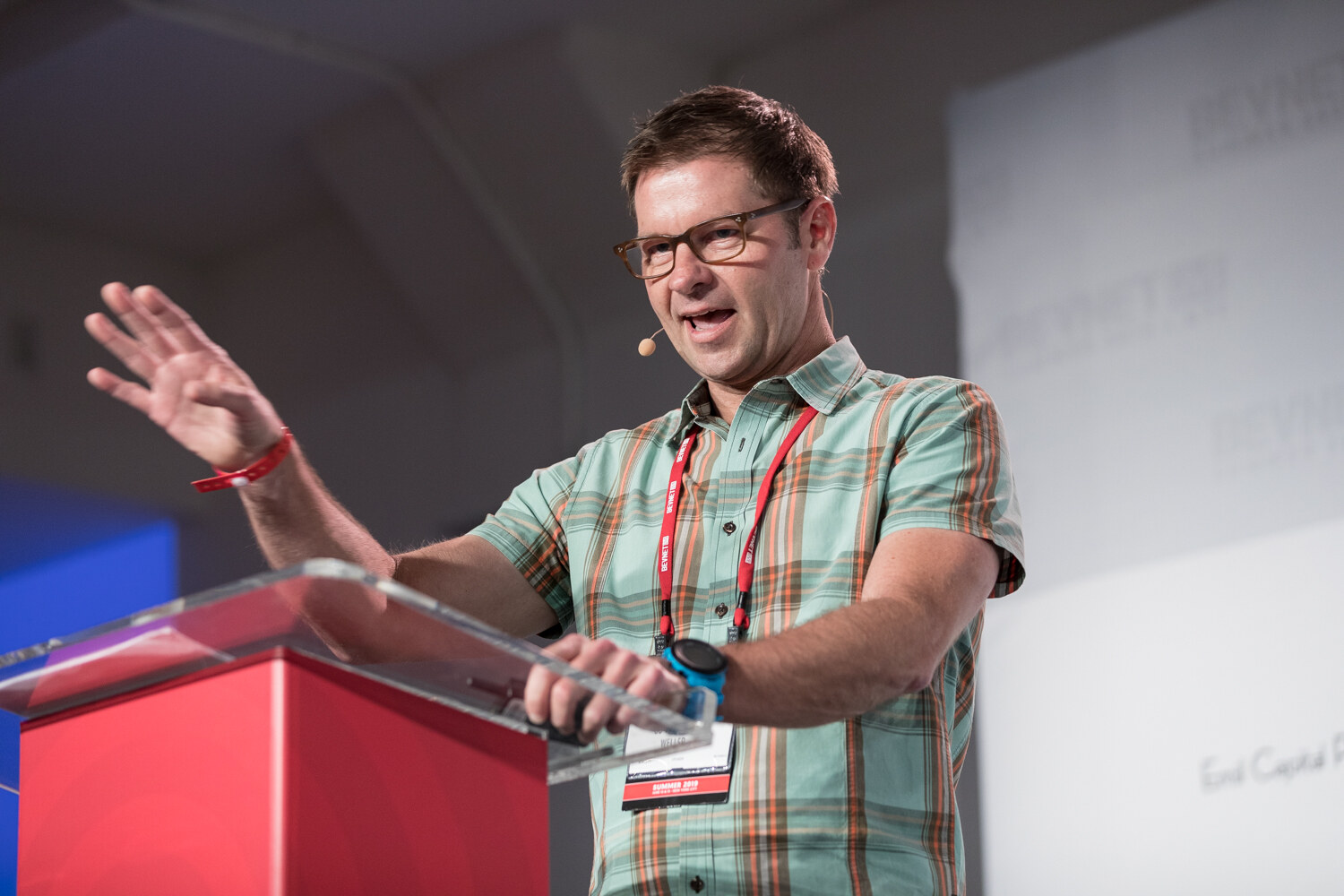 Man in a mint green plaid shirt with a raised hand at the red podium.