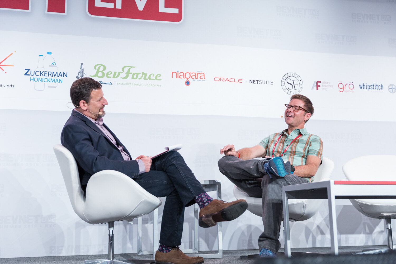 Interviewer in a navy blazer and a man in a mint green plaid shirt seated in white chairs.