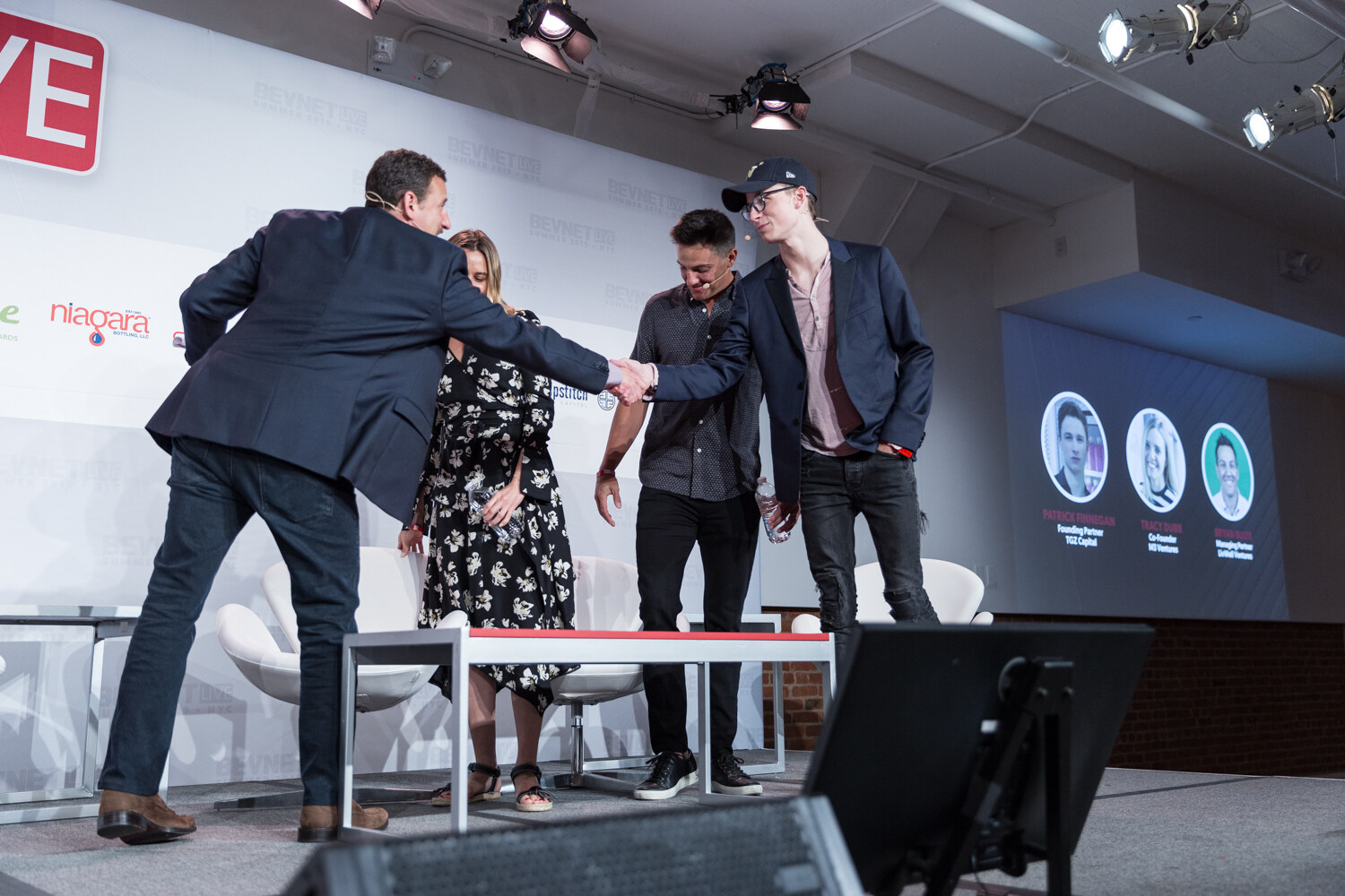 Moderator shaking hands with panelists on stage as a screen behind them shows their portraits.