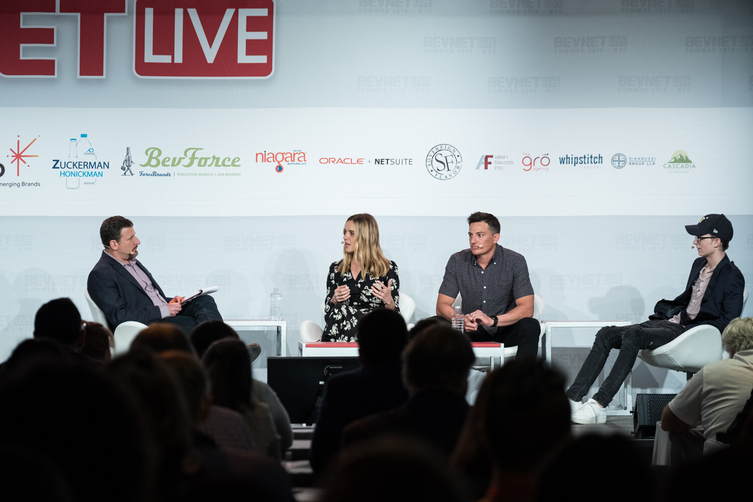 Moderator in a navy jacket and three panelists seated in white chairs on the BevNET Live stage, with the audience in silhouette in the foreground.