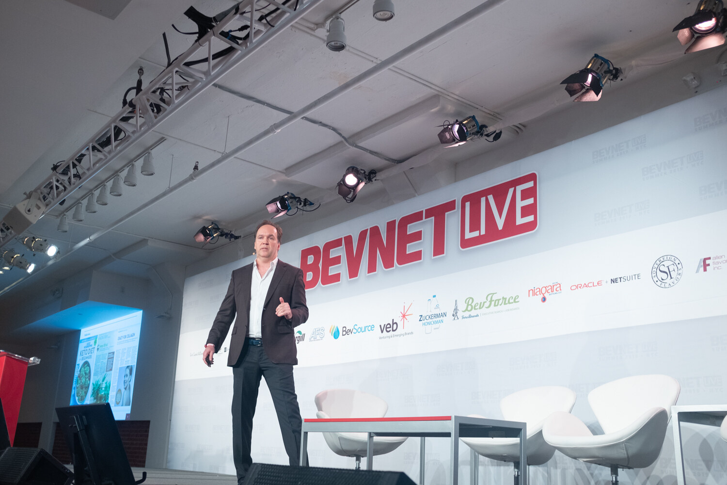 Speaker in a dark blazer and white open-collared shirt walking the stage in front of the BevNET Live sign, with a slide glowing at left.