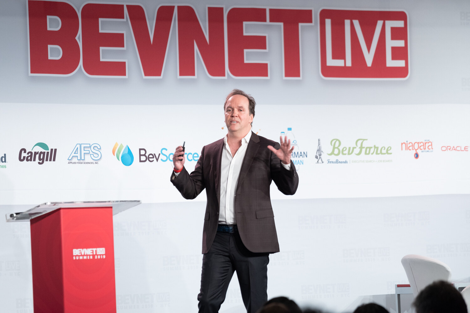 Speaker in a dark blazer gesturing near the red podium in front of the BevNET Live sign and sponsor logos.