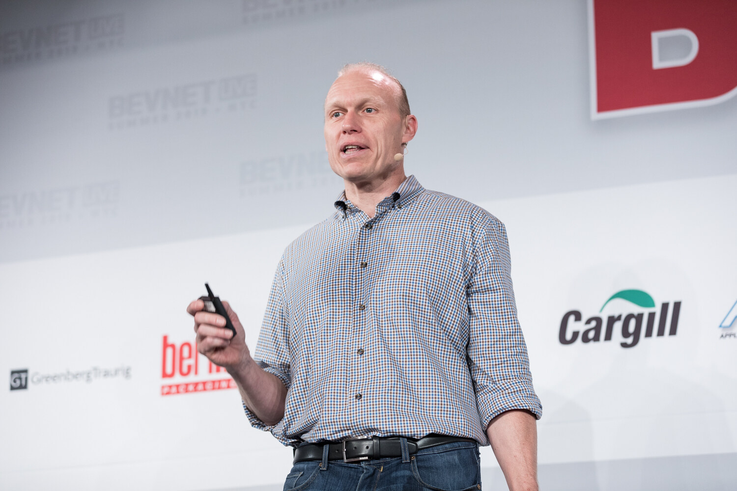 Man in a gray checked shirt holding a clicker and a blue bottle while speaking on stage in front of Berlin Packaging and Cargill logos.
