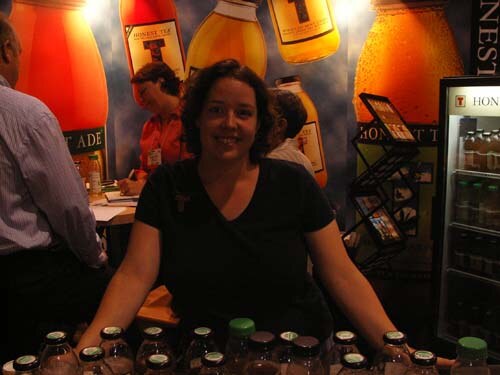 A smiling woman with dark curly hair in a black shirt stands behind a counter of bottles in the Honest Tea booth; other people stand behind her, with large bottle graphics and a cooler nearby.