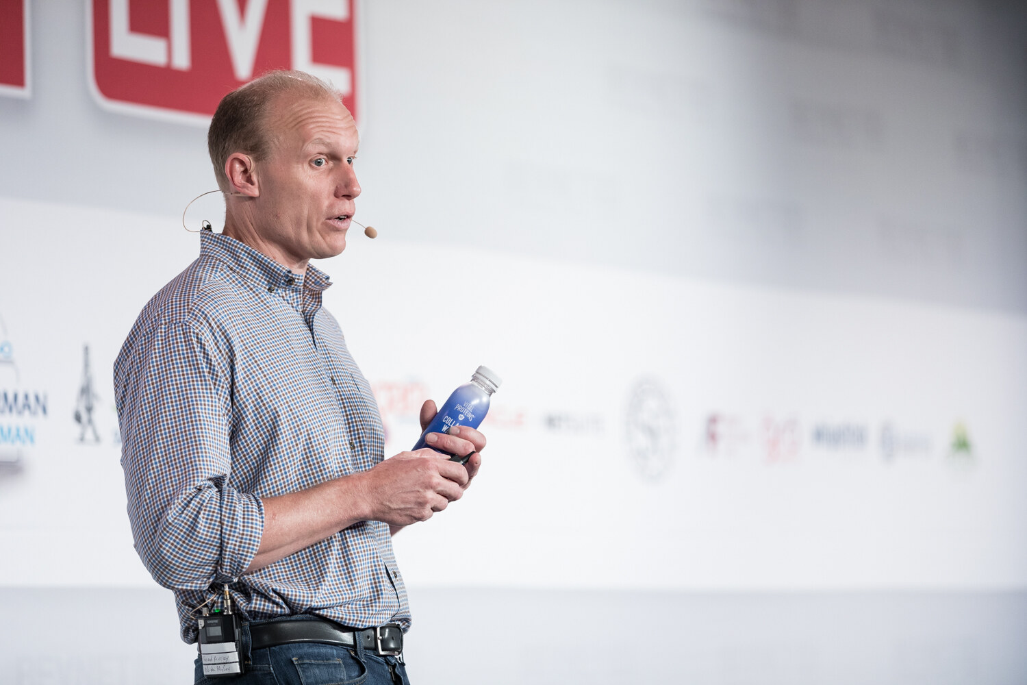 Speaker holding a blue bottle and clicker while speaking into a headset microphone.