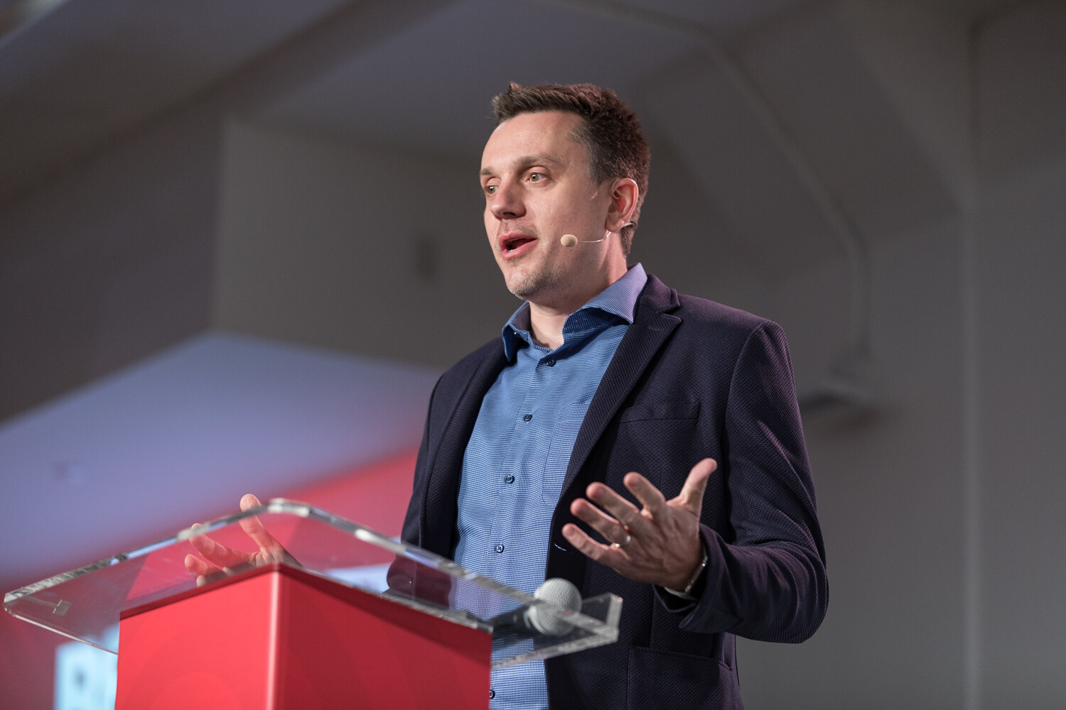 Man in a navy blazer and blue shirt gesturing with an open hand at the red podium.