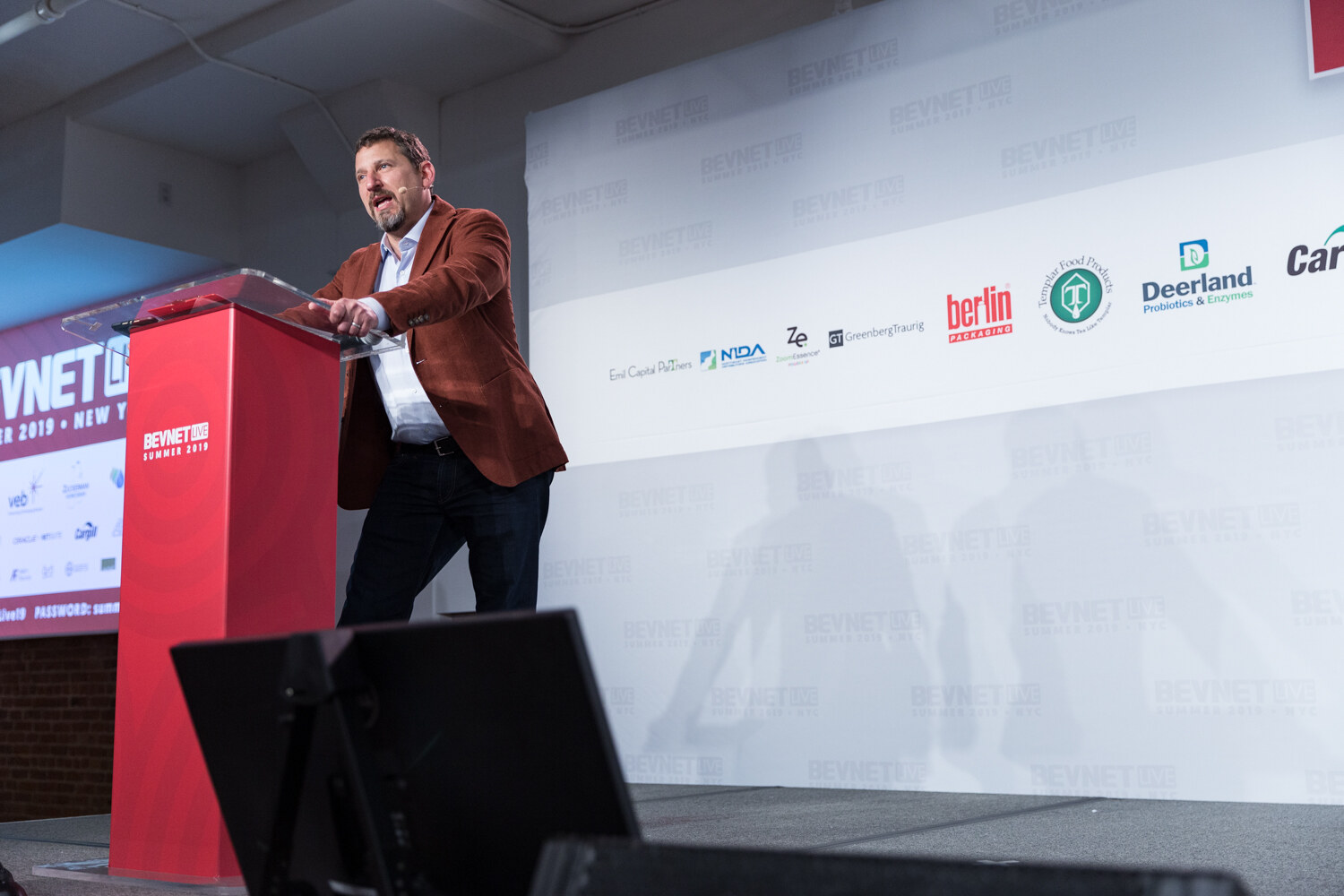 Host in a rust-colored blazer leaning on the red podium beside a screen that reads BevNET Summer 2019 New York.