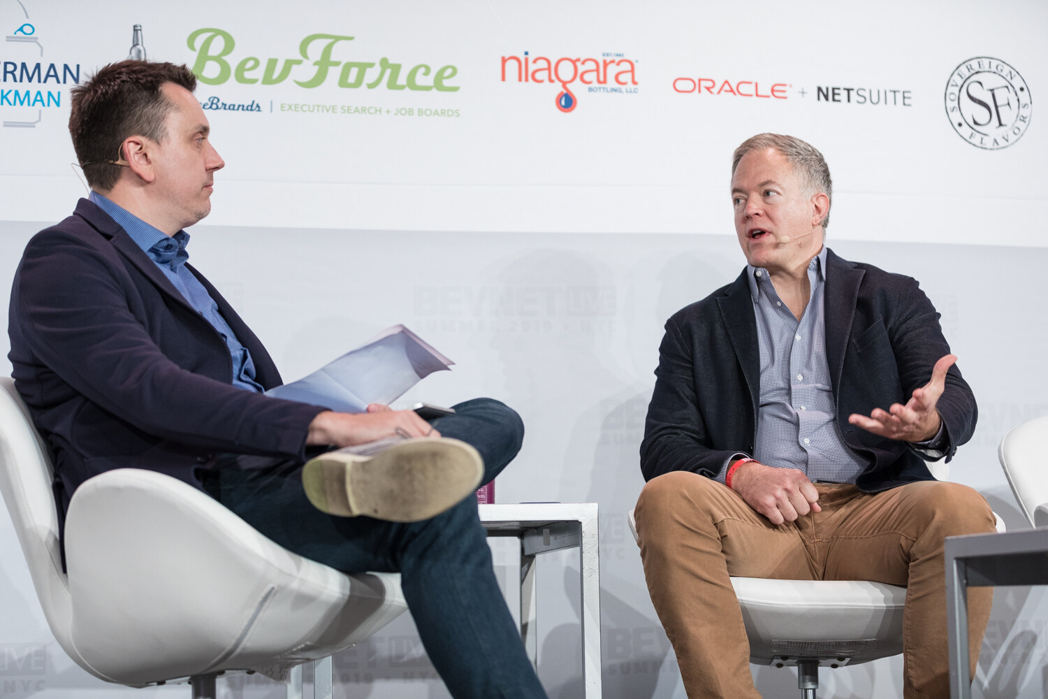 Interviewer holding papers and the man in a navy blazer talking in white chairs in front of BevForce and Niagara logos.