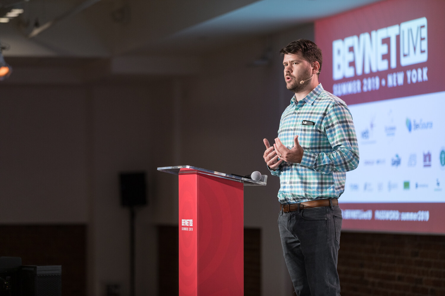 Speaker in a teal checked shirt at the red podium with a BevNET Live Summer 2019 New York slide behind him.