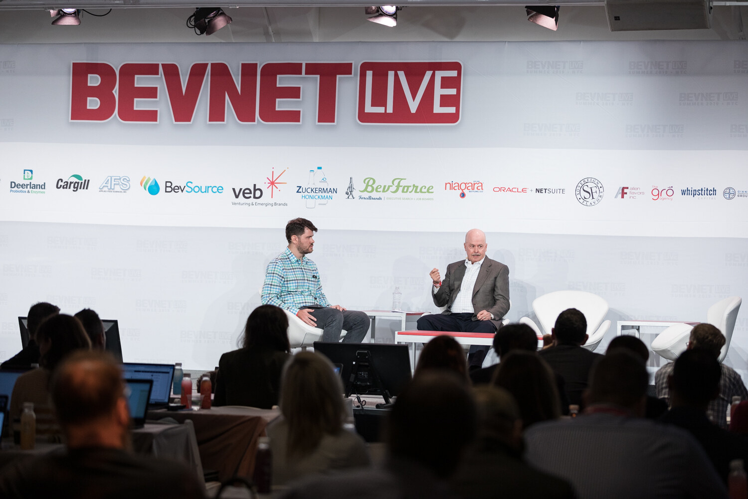 Wide view of a two-person fireside chat on stage with the audience in the foreground.