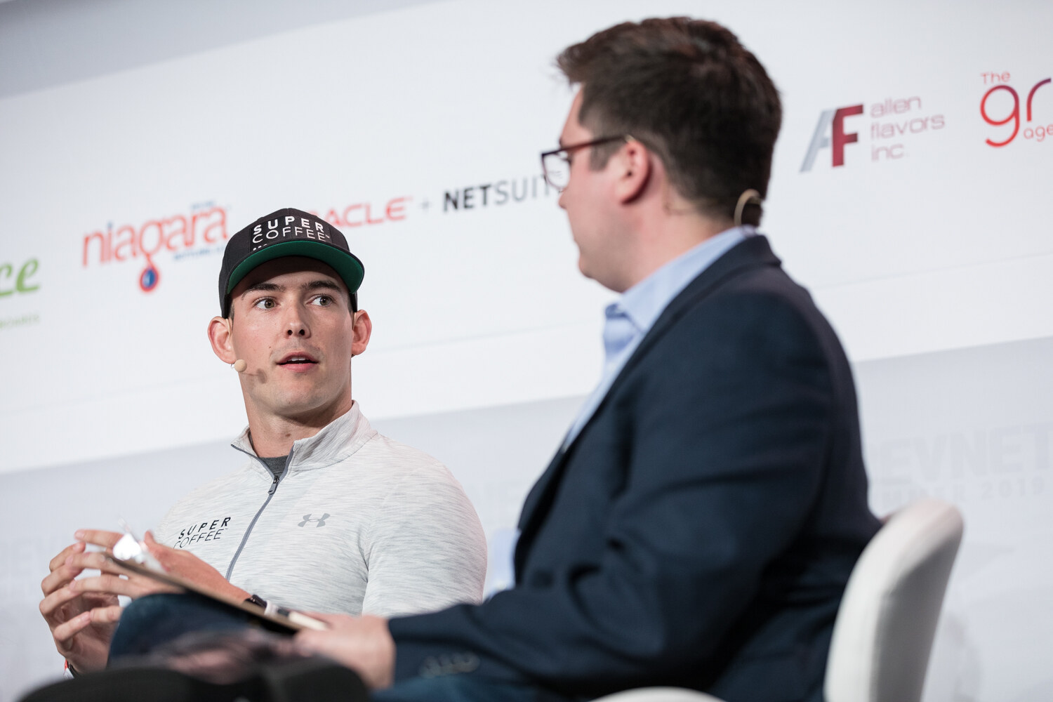 Man in a Super Coffee cap and gray pullover speaking with a clipboard in his lap, the moderator in a navy blazer at right.