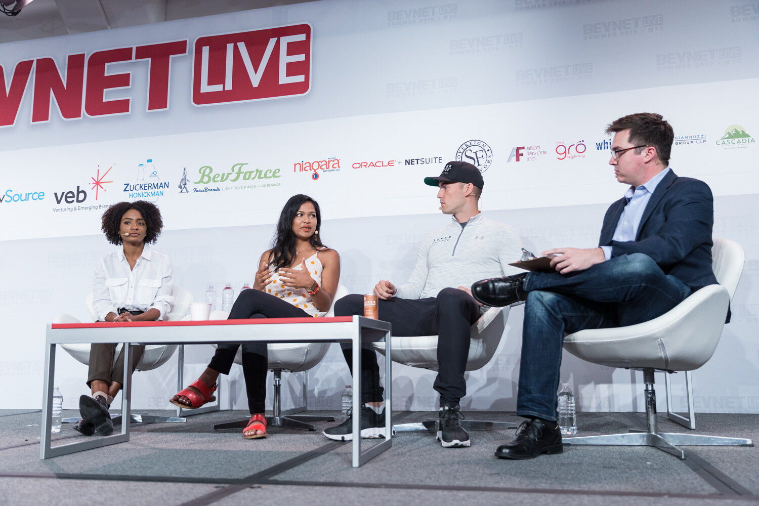 Four people seated on stage around a low red table, the moderator in a navy blazer at right holding a clipboard.